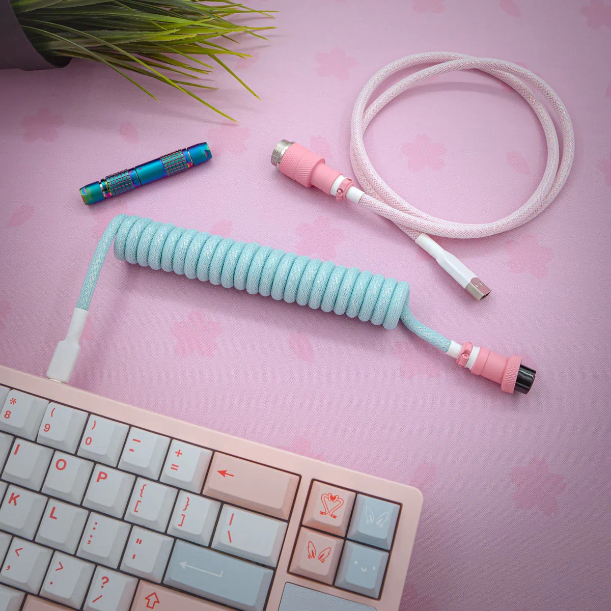 Blue pink sparkle coiled cable pbt noel A pastel desk setup with a white and pink mechanical keyboard, a Blue Pink Sparkle Coiled Cable PBT Noel, a pink braided USB cable, a colorful flashlight, and a plant on a cherry blossom-patterned desk mat.