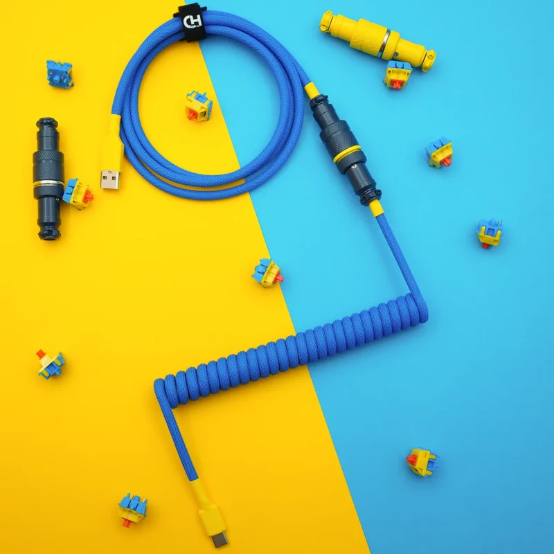 Blau Gelb Spiralkabel USB C Overhead Ein leuchtend blaues Spiral-Tastaturkabel mit USB-C und abnehmbaren Pilotenanschlüssen, umgeben von bunten Tastenschaltern. Ideal für benutzerdefinierte Setups.
