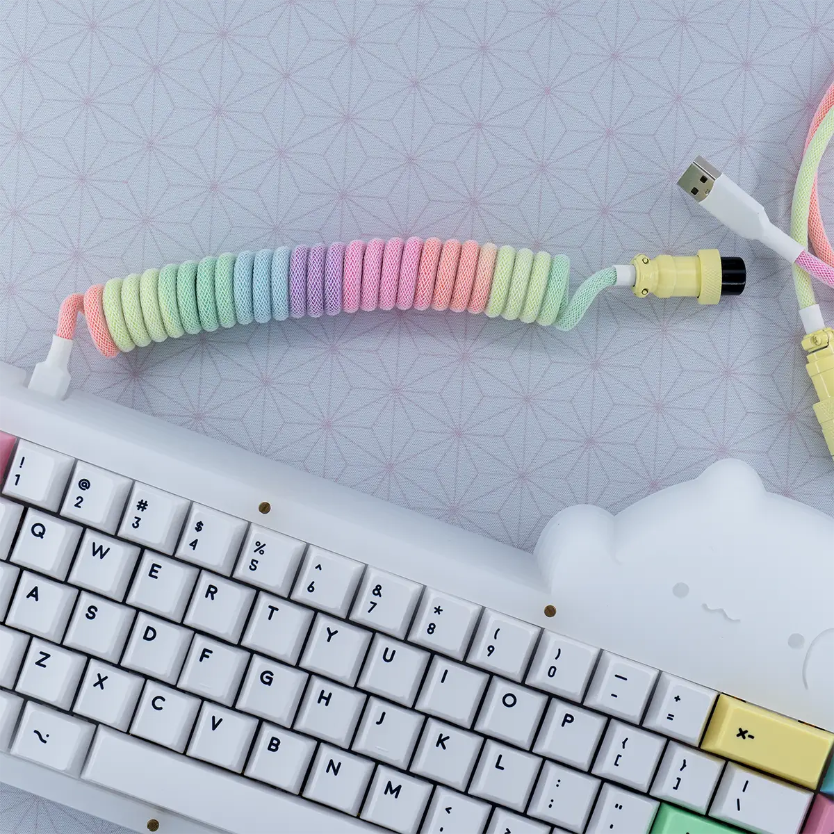 Bongo65 keyboard pbt milkshake coiled keyboard cable The Bongo65 keyboard with pastel PBT milkshake keycaps sits on a patterned surface, next to a rainbow pastel Bongo65 coiled keyboard cable featuring yellow connectors.
