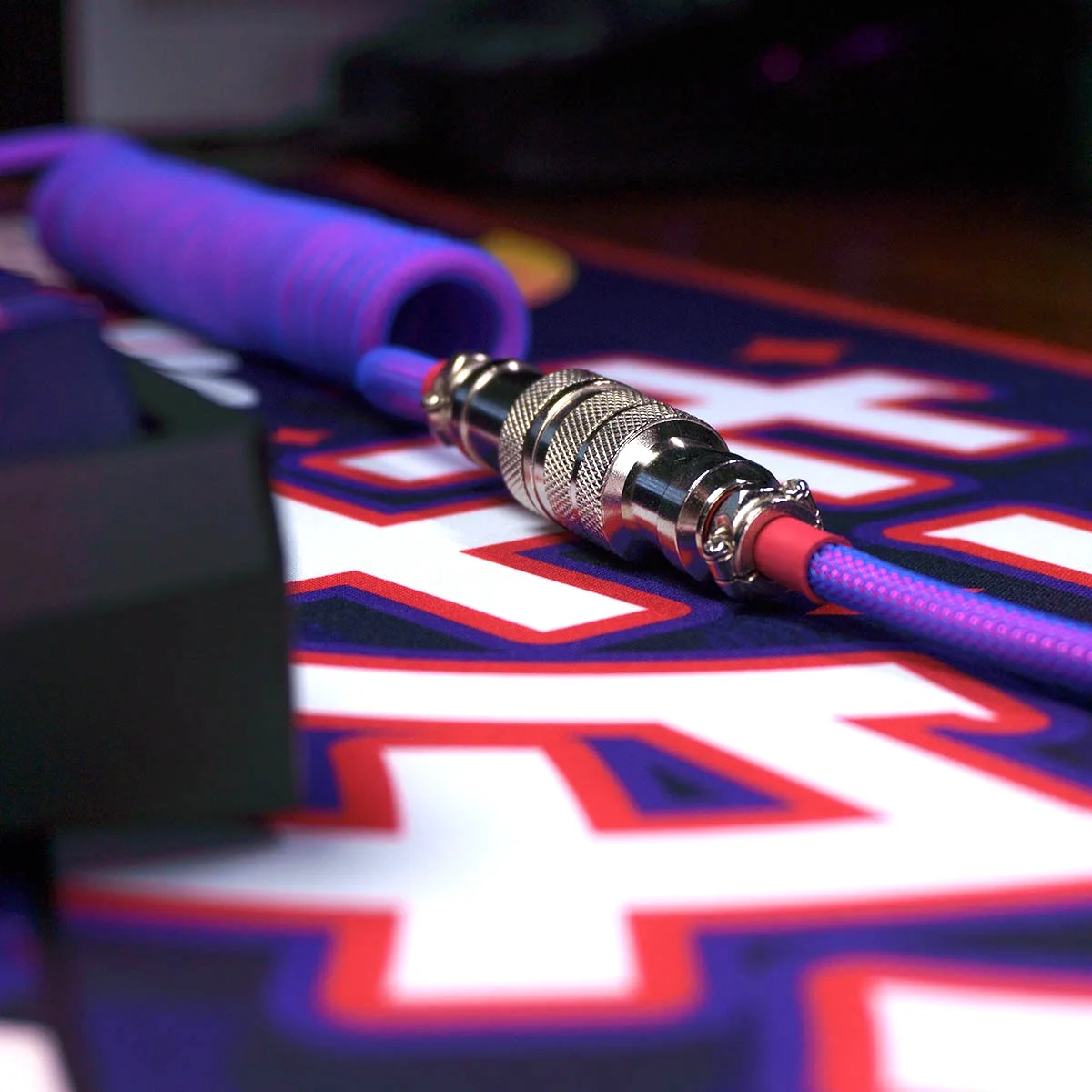Coiled cable laser themed with gx16 silver 4 pins A close-up view of a laser-themed coiled purple cable with a GX16 silver 4-pin connector, lying on a colorful surface with bold text and graphics.