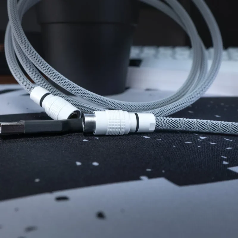 Custom straight black and white keyboard cable yc8.webp A coiled, black & white USB cable with yc8 connector white and sleek connectors rests on a black and gray patterned mouse pad, with a black cup and keyboard blurred in the background.