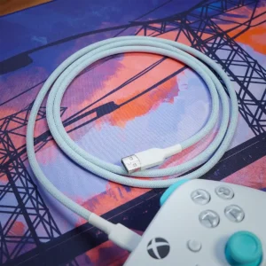 Câble bleu droit personnalisé pour contrôleur Un câble bleu droit personnalisé est enroulé sur un tapis de souris coloré avec un motif abstrait représentant une ligne électrique, relié à une manette de jeu blanche avec des boutons turquoise.