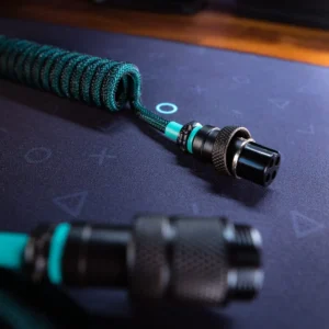 A close-up of the Gmk Hammerhead coiled cable for keyboard, featuring teal and black sleeving, metal connectors, and detachable design, resting on a dark mouse pad with geometric symbols.