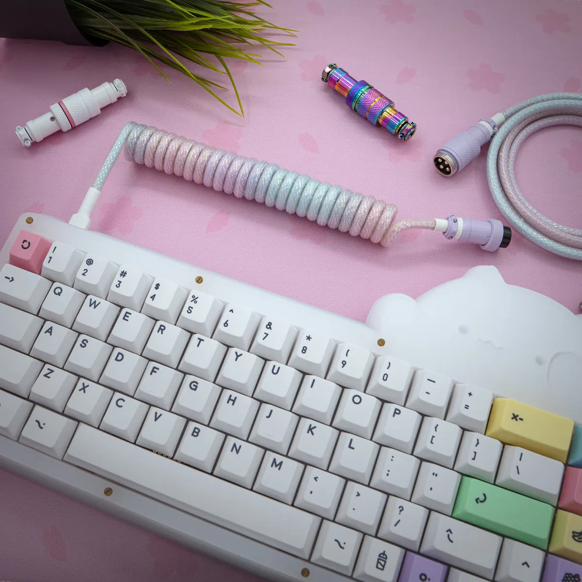 Kawaii sparkle coiled cable A pastel mechanical keyboard with multicolored keycaps sits on a pink desk mat. The Kawaii sparkle coiled cable with colorful connectors is nearby, and a small plant appears in the background.