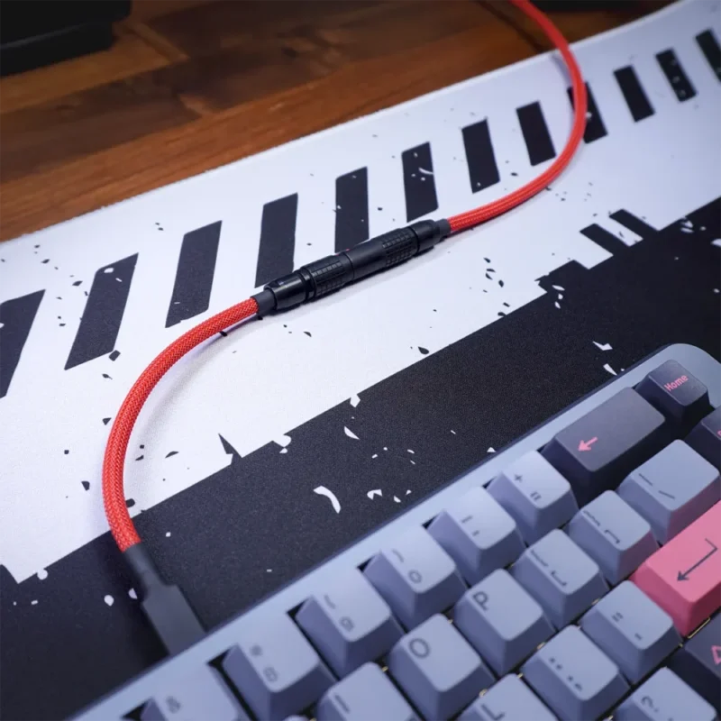 Nahaufnahme einer mechanischen Tastatur mit grauen und rosa Tasten auf einer schwarz-weißen Schreibtischunterlage. Über der Tastatur verläuft ein rotes gerades Tastaturkabel mit schwarzen Lemo-Steckern.