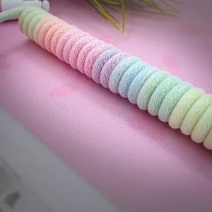 Câble enroulé de couleur arc-en-ciel scintillant Le câble spiralé Sparkle rainbow, enveloppé dans une maille pastel, repose sur une surface rose avec de subtils motifs en forme de cœur et des feuilles de plantes vert tendre en arrière-plan.