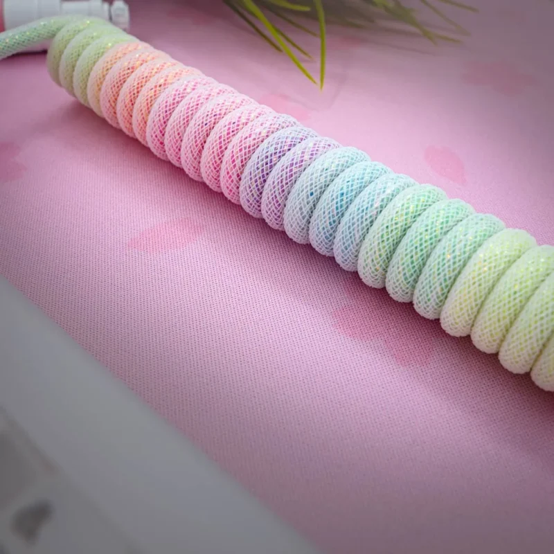 Sparkle rainbow coiled cable The Sparkle rainbow coiled cable, wrapped in pastel mesh, rests on a pink surface with subtle heart shapes and soft green plant leaves in the background.