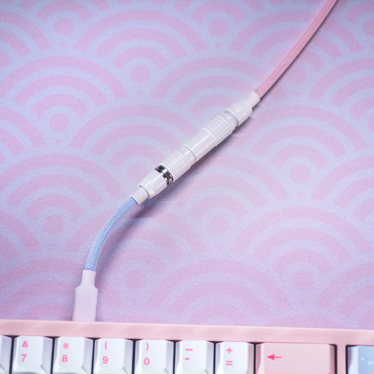 Straight blue and pink keyboard cable A Custom Straight Keyboard Cable - Detachable with a white connector is plugged into a pastel pink keyboard on a pink and purple wave-patterned desk mat.